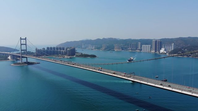 Long suspension bridge span over channel, aerial camera soar towards impressive structure. Part of Lantau Link, Tsing Ma bridge carries mixed auto and rail traffic on two decks. Cars ride on highway