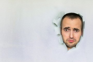 Sad guy making hole in paper, looking down with downcast eyes, pursing his lips, blue background, copy space, for advertising