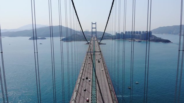 Impressive lengthwise aerial perspective of large suspension bridge, camera fly along road, between cables. Light traffic on highway, modern Lantau Link at Hong Kong city. Bridge span across channel