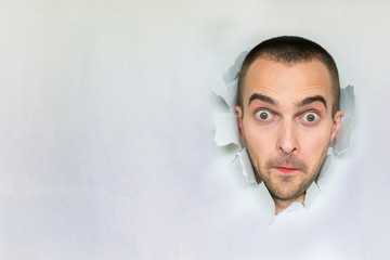 Amazed man making hole in paper, looking into the camera, pursing her lips, blue background with a...