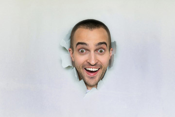 Happy young guy making hole in paper, looking at the camera, blue background, copy space, for...