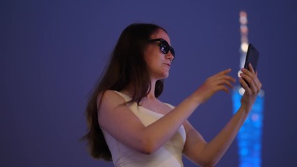 Woman gesticulate and speak, share emotions during video call to friend, portrait shot at Huacheng Plaza. Lady stay against blurred Canton tower with colorful glowing facade at evening twilight.