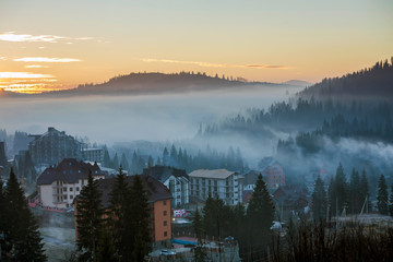 Resort village houses buildings on background of foggy blue mountain hills covered with dense misty spruce forest under bright pink sky at sunrise. Mountain landscape at dawn.