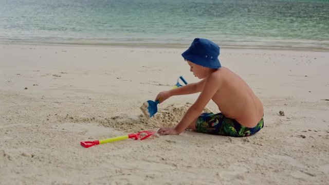 A little boy in shorts and a blue hat, sits on the beach near the water and digs a hole with a shovel.