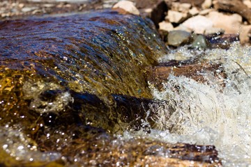 Clean river water flows over the stones and envelops them. Bright sun rays are reflected in the flowing water and betray its brilliance. Water hits the stones forming waves and splashes.
