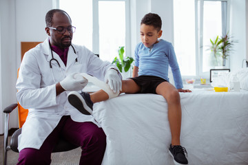 Dark-skinned pediatrician putting bandage on leg of little boy