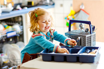 Cute little toddler girl helping in the kitchen with dish washing machine. Happy healthy blonde...