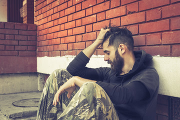 Homeless man drug and alcohol addict sitting alone and depressed on the street leaning against a red brick building wall feeling anxious and lonely in the morning, social documentary concept  - obrazy, fototapety, plakaty