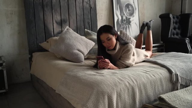 Young Asian woman in bed messaging with mobile telephone at home. A girl with dark skin lies in bed with