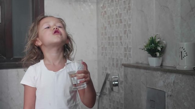 Cute little curly child girl rinses her mouth with water and spits water into the sink. Portrait shot in mirror reflection.