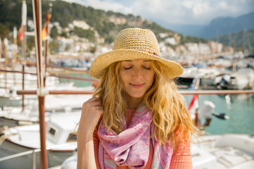 Portrait of young woman with light curly hair in straw hat enjoying sun and breeze, smiling, eyes closed. Sunny harbor with boats and yachts, green mountains on background. Enjoying life, traveling,