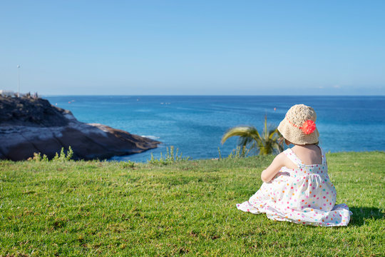 Adorable Caucasian little girl wearing a bright light summery dress and a straw hat with a large flower, resting on the neatly cut grass and looking towards the coastal landscape in front of her