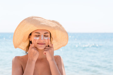 Beautiful young woman in hat is applying sunblock under her eyes and on her nose like Indian. Sun...