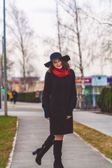 The happy girl with smile walks around the city in black coat and hat with wide fields. Red lipstick and scarf. Fashion, style.