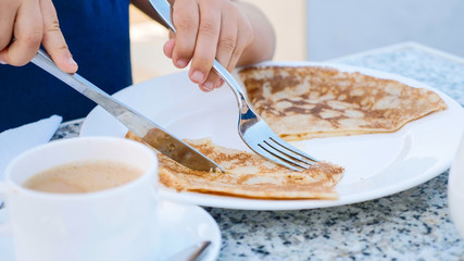 Little girl is cutting a pancake on a plate with knife and fork for breakfast.