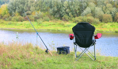 sports summer fishing with one rod and comfortable folding tourist chair on river bank among green grass and beautiful trees