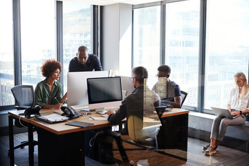 Young creative team working together at computers in a casual office, seen through glass wall
