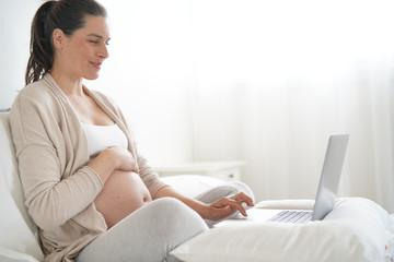 Naklejka premium Pregnant woman relaxing on bed, using laptop