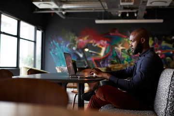 Mid adult black male creative sitting at table in office dining area using a laptop, side view