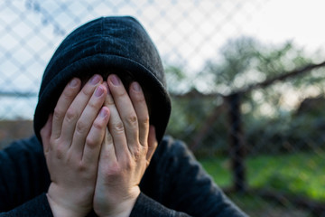 Frustrated teenage boy covering hes face in front of correctional institutes wired fence,...