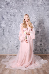 Fototapeta na wymiar Young beautiful woman with blond wavy hairstyle wearing tender pink wedding dress. Fragile bride portrait in full length maxidress. Close up, copy space, background.