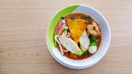 Noodle bowl that consist of fish ball, vegetable, soup and more placed on the white cup and disk on the wooden table.