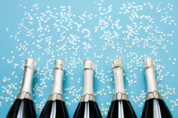 Five Champagne bottles with holographic confetti stars on light blue background. Copy space, top view