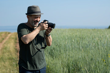 Adult man in a hat holding a camera and laughs. The background of the image is a country road among a field of wheat. Copy space. Concept: the fun of the hobby.