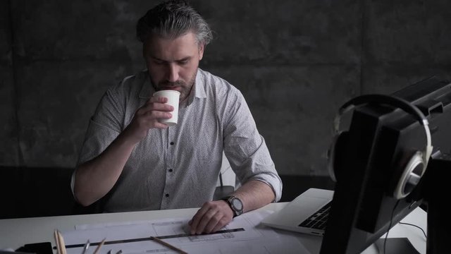 middle-aged man drinks coffee and continues to work with drawings and documents in the office.