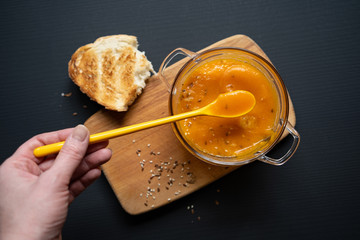 a young girl's hand is holding a yellow spoon submerged in pumpkin soup, a wooden board on a black background, a piece of bread, sesame and flax seeds