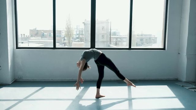 Young female performs a backflip with hands in the dance stodio Flexible woman doing splits in fashion look. View on window sunny day