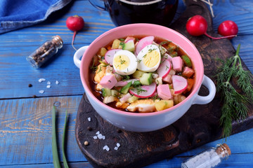 Russian cold soup Okroshka with vegetables, sausage, eggs and kvass in a ceramic bowl on the blue table