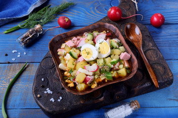 Russian cold soup Okroshka with vegetables, sausage, eggs and kvass in a bowl on the blue table