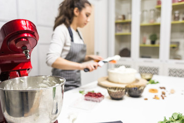 In the foreground is a metal bowl with a whipped cream and a red electric mixer. In the background, the confectioner smoothes white cream on a biscuit cake with a culinary spatula.