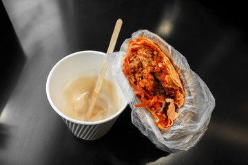 vegetarian shawarma with falafel in plastic package and a cup of coffe with wooden spoon on the dark glassy table, view from above. Falafel as part of vegetarian cuisine, and as a form of street food