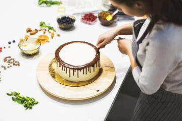 A confectioner squeezes liquid chocolate from a pastry bag onto a white cream biscuit cake on a wooden stand. The concept of homemade pastry, cooking cakes.