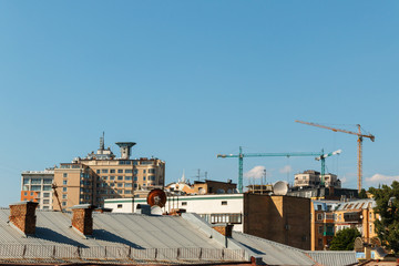 Fototapeta premium cityscape with roofs of houses and three construction cranes on the blue sky background with space for text. Turret slewing cranes, old and new architecture in Kyiv.