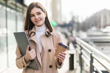 Portrait of joyful european woman walking against office building outdoor with silver laptop and takeaway coffee in hands