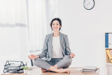 beautiful smiling businesswoman in suit sitting on desk and meditating in Lotus Pose in office