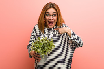 Young woman holding a plant surprised pointing at herself, smiling broadly.