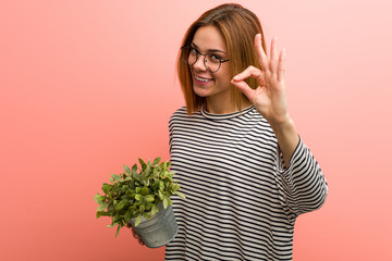 Young woman holding a plant cheerful and confident showing ok gesture.