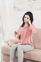 sad woman sitting on couch with glass of water and touching face at home