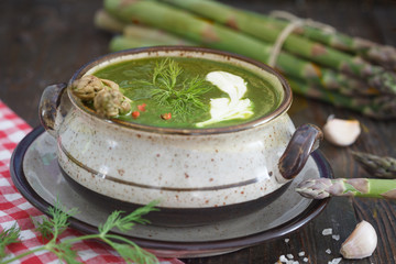 Creamy asparagus soup decorated with cream, dill and red pepper. Green asparagus in background, wooden table.