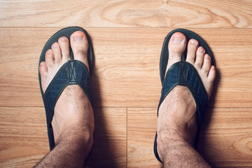 Male hairy feet wearing blue flip flops standing on a brown wooden floor - Powered by Adobe