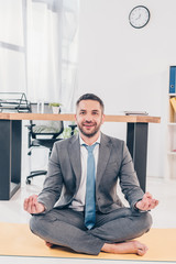 handsome smiling businessman meditating in Lotus Pose on fitness mat in office