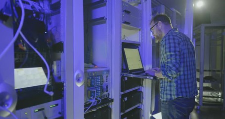 Network administrator in glasses in server room types text on laptop - Powered by Adobe