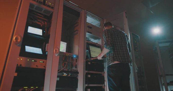 Side view of young male programmer in eyeglasses connecting wires in server cabinet while working with mainframe in data center