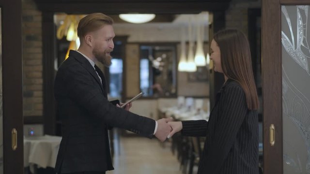 Male and female business partners meet in the restaurant. People shaking hands. Lady in an elegant suit coming to the bearded man waiting for her near the entrance