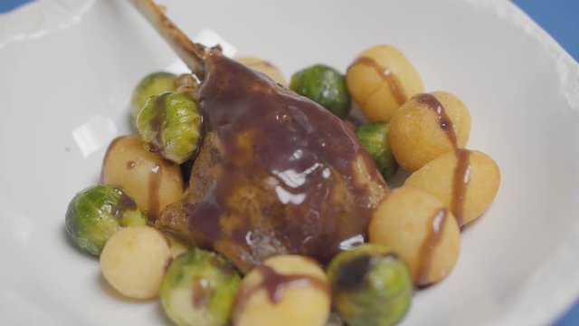 White plate with duck leg, potato balls and brussels sprouts on the sause close up.