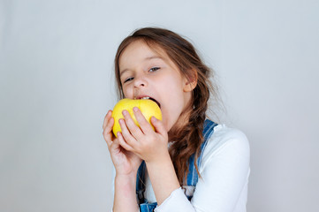 Emotional portrait little beautiful girl with pigtails in jeans overalls eating bites holding an apple. 6-7 years studio